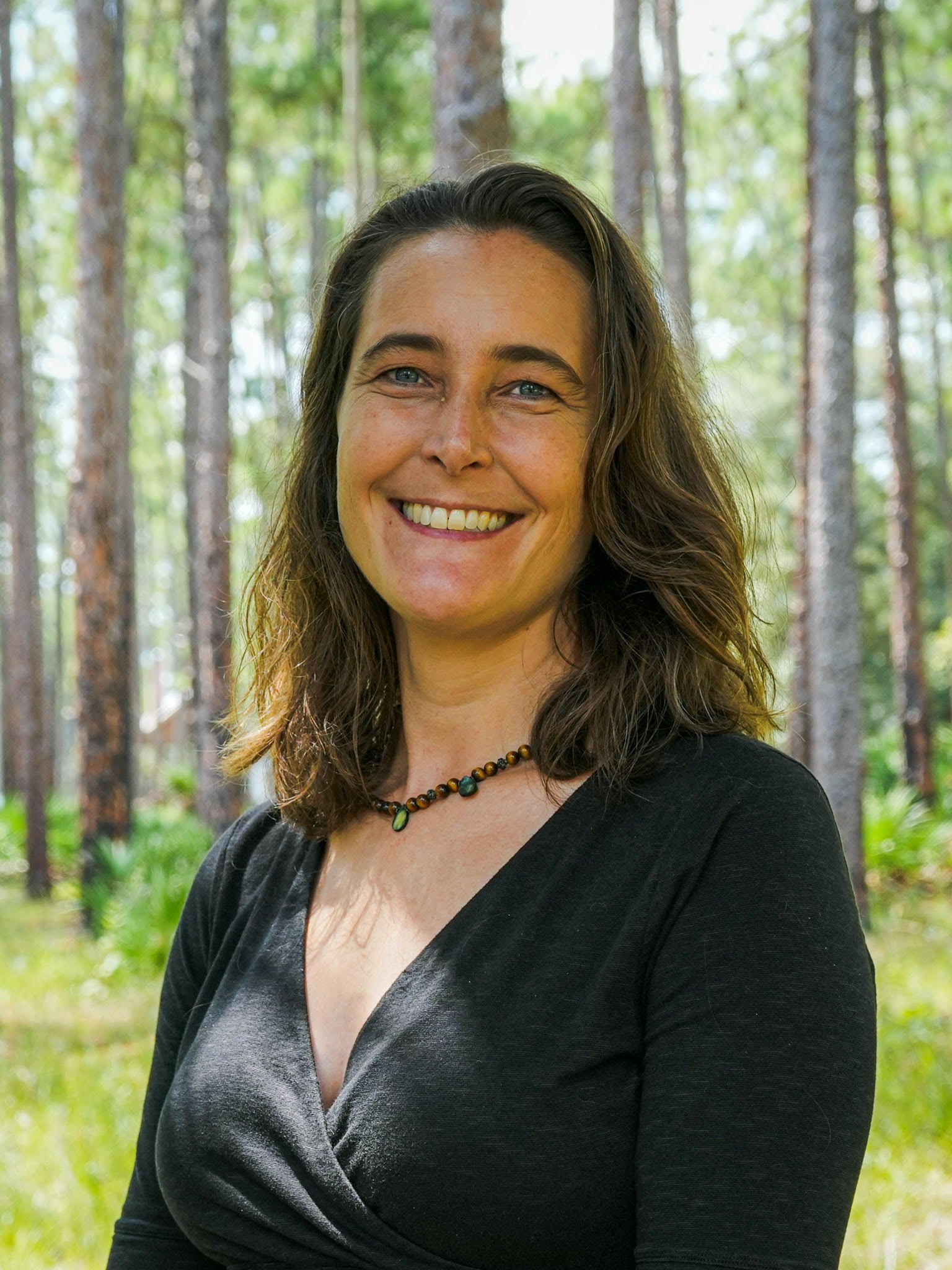 Geraldine is a white woman with shoulderlength brown wavy hair. She is wearing a black top and a necklace from small brown and green stones. The backdrop is a Florida pine forest